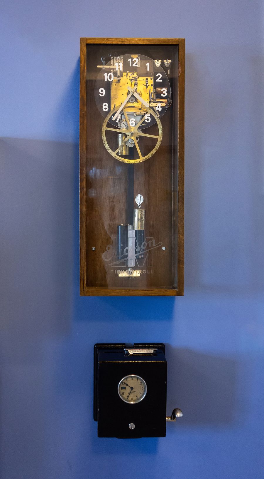 A wooden master clock with brass gears above a black punch-card time recorder mounted on a blue wall