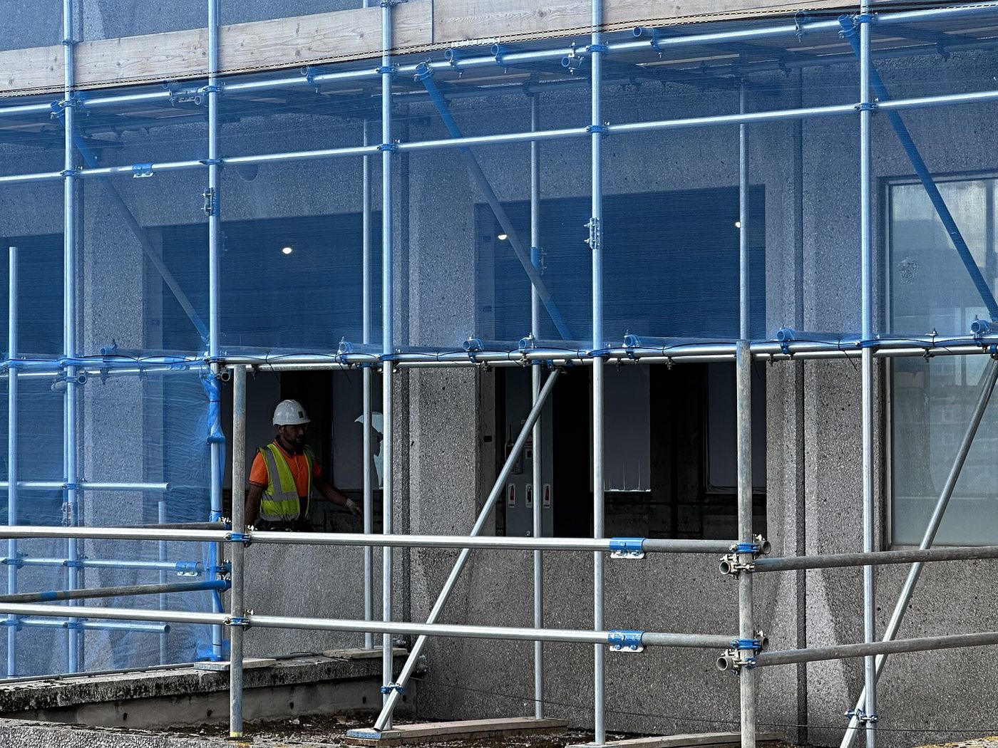 A builder in a white helmet and hi-vis vest walks behind blue scaffolding on a concrete building